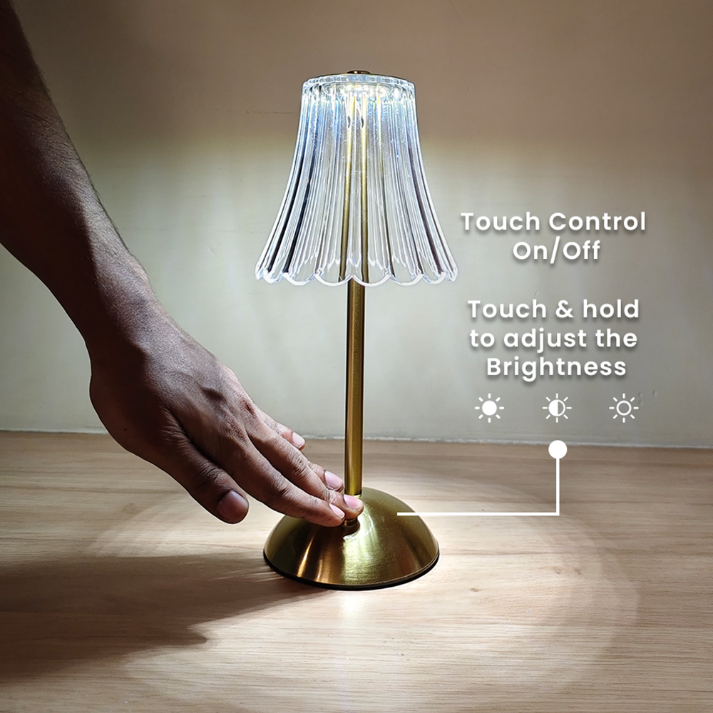 Gold table lamp with clear lampshade on a wooden surface, hand touching the base.
