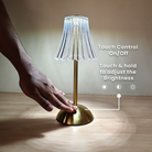 Gold table lamp with clear lampshade on a wooden surface, hand touching the base.